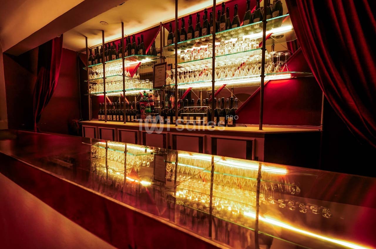 Bar élégant avec comptoir brillant, étagères illuminées contenant bouteilles et verres, décor rouge et rideaux.