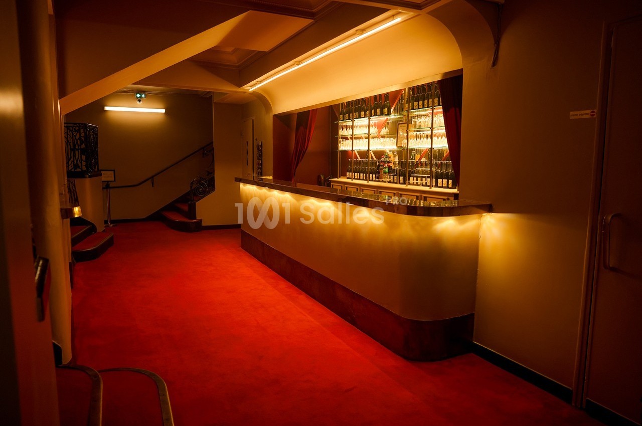 Bar éclairé avec des étagères de bouteilles, situé dans un couloir à moquette rouge et éclairage tamisé.