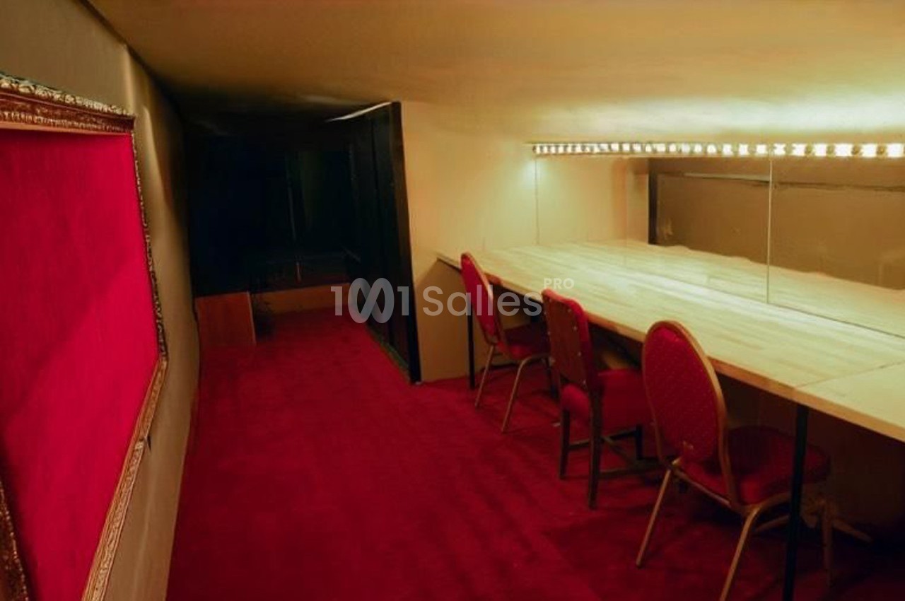 Salle avec moquette rouge, table en bois clair, chaises rouges et miroir éclairé par des ampoules.