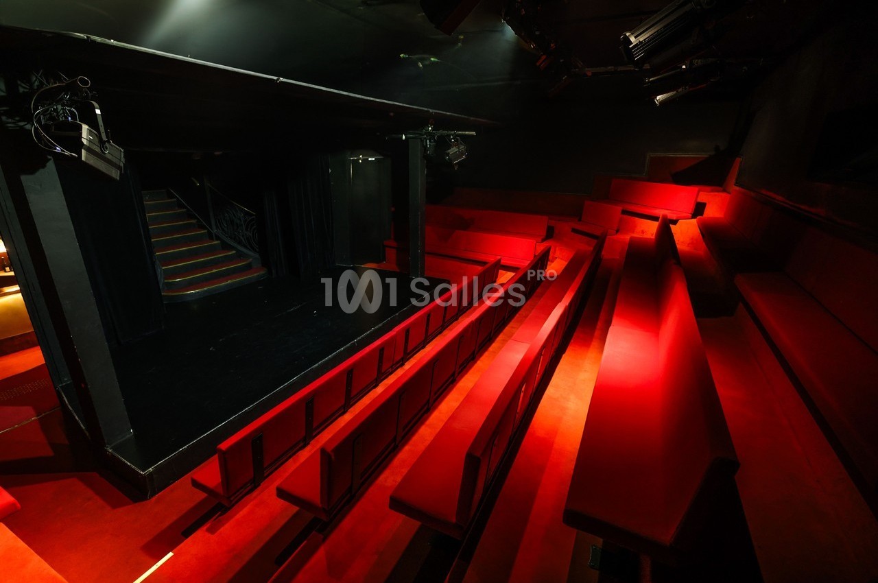 Salle de théâtre sombre avec des sièges rouges en gradins et une petite scène noire éclairée.