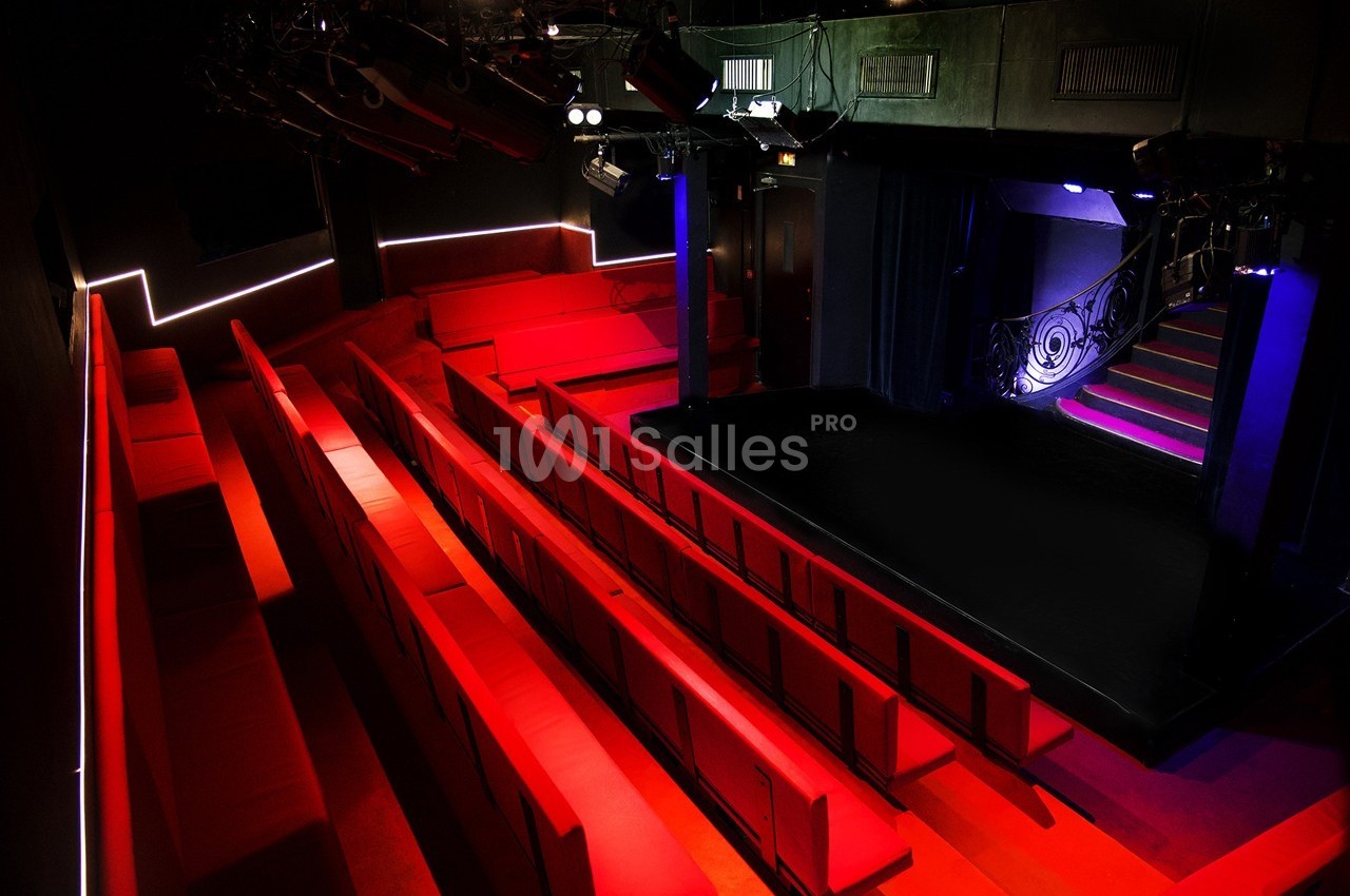 Salle de spectacle vide avec des sièges rouges, une scène éclairée et un escalier sur le côté.
