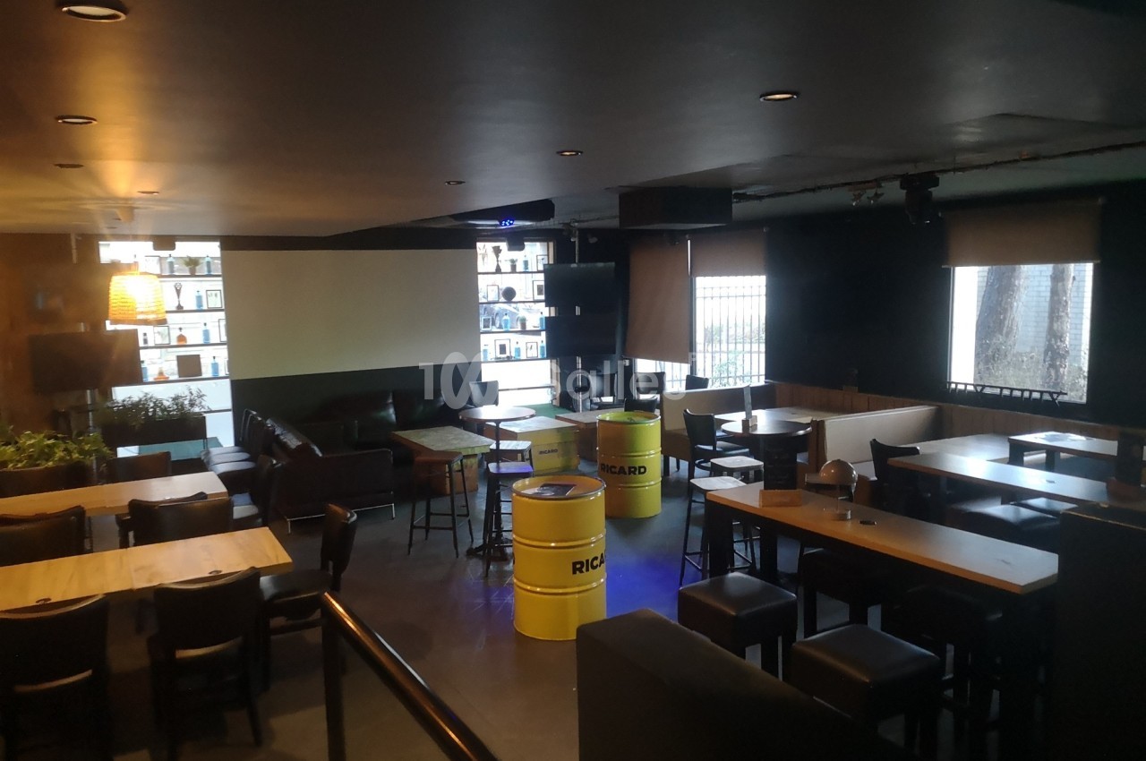 Salle de bar avec tables, chaises, tonneaux jaunes en guise de tables et étagères éclairées en arrière-plan.