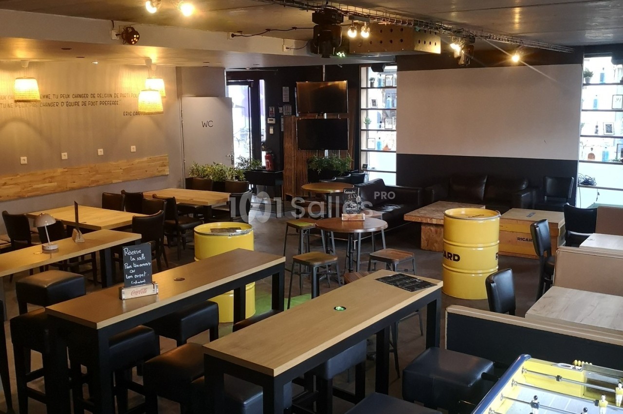 Salle de bar moderne avec tables en bois, fûts recyclés comme mobilier, canapé, baby-foot et éclairage tamisé.