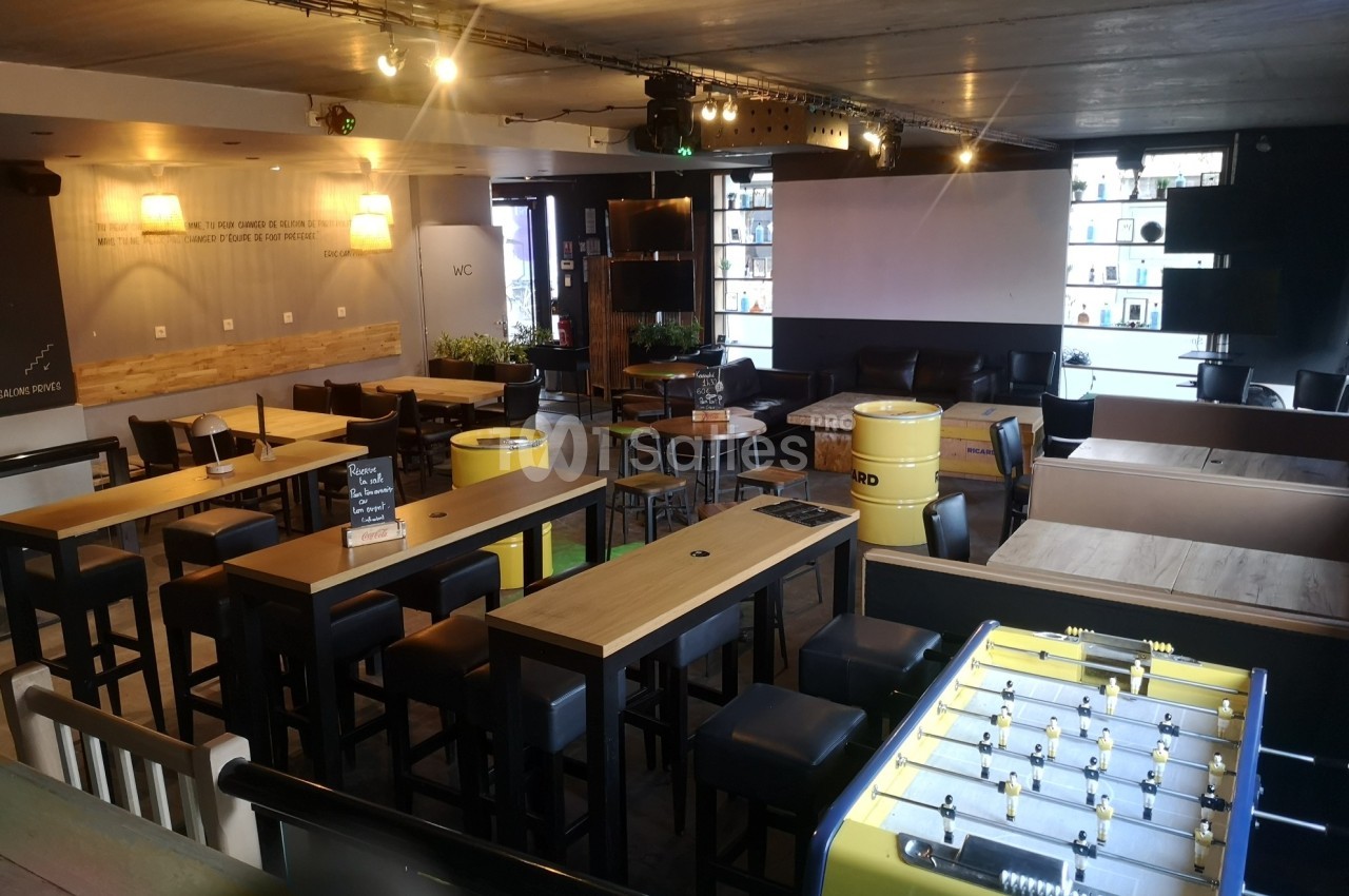Salle de bar moderne avec tables hautes, tabourets, baby-foot jaune et éclairage chaleureux.