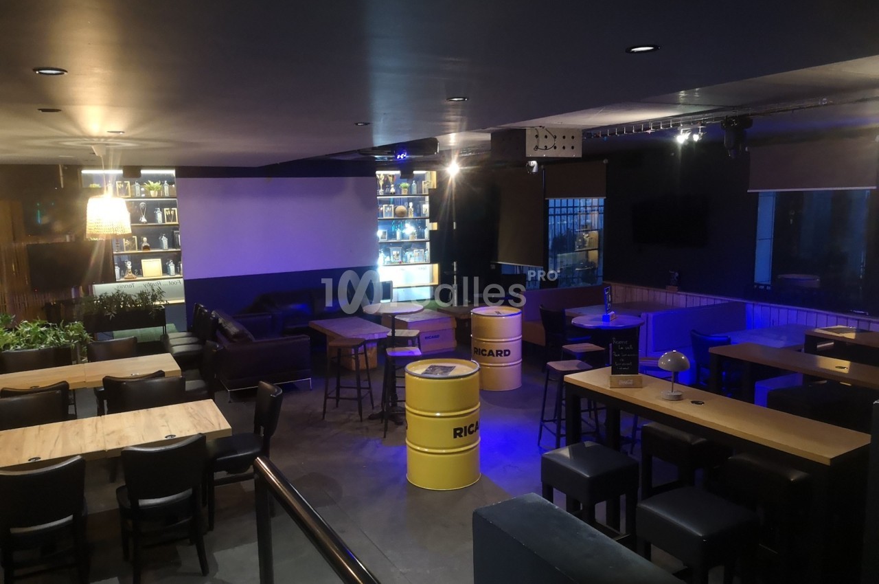 Salle de bar avec tables en bois, tonneaux jaunes en guise de tables, éclairage tamisé et étagères illuminées.