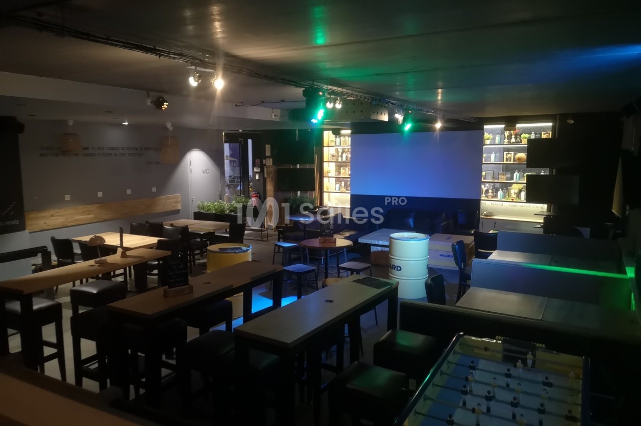 Salle de bar avec tables hautes, baby-foot, éclairage tamisé et écran géant au fond.