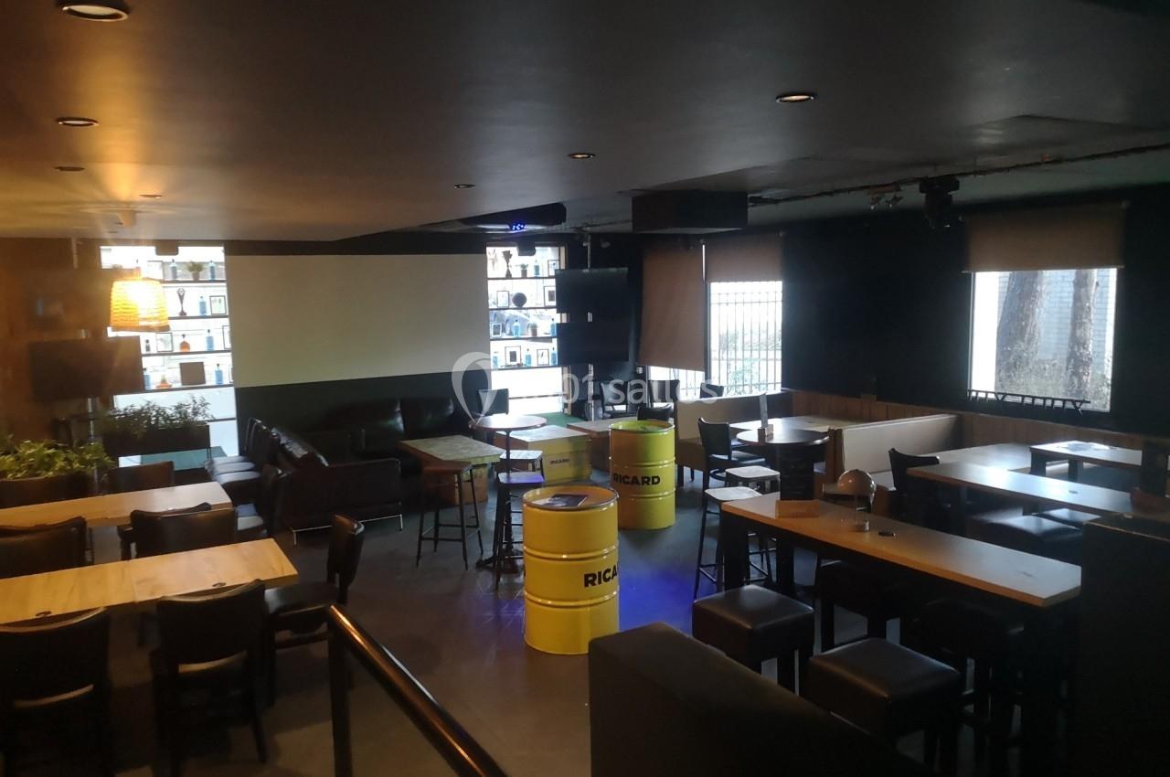 Salle de bar avec tables, chaises, tonneaux jaunes en guise de tables et étagères éclairées en arrière-plan.