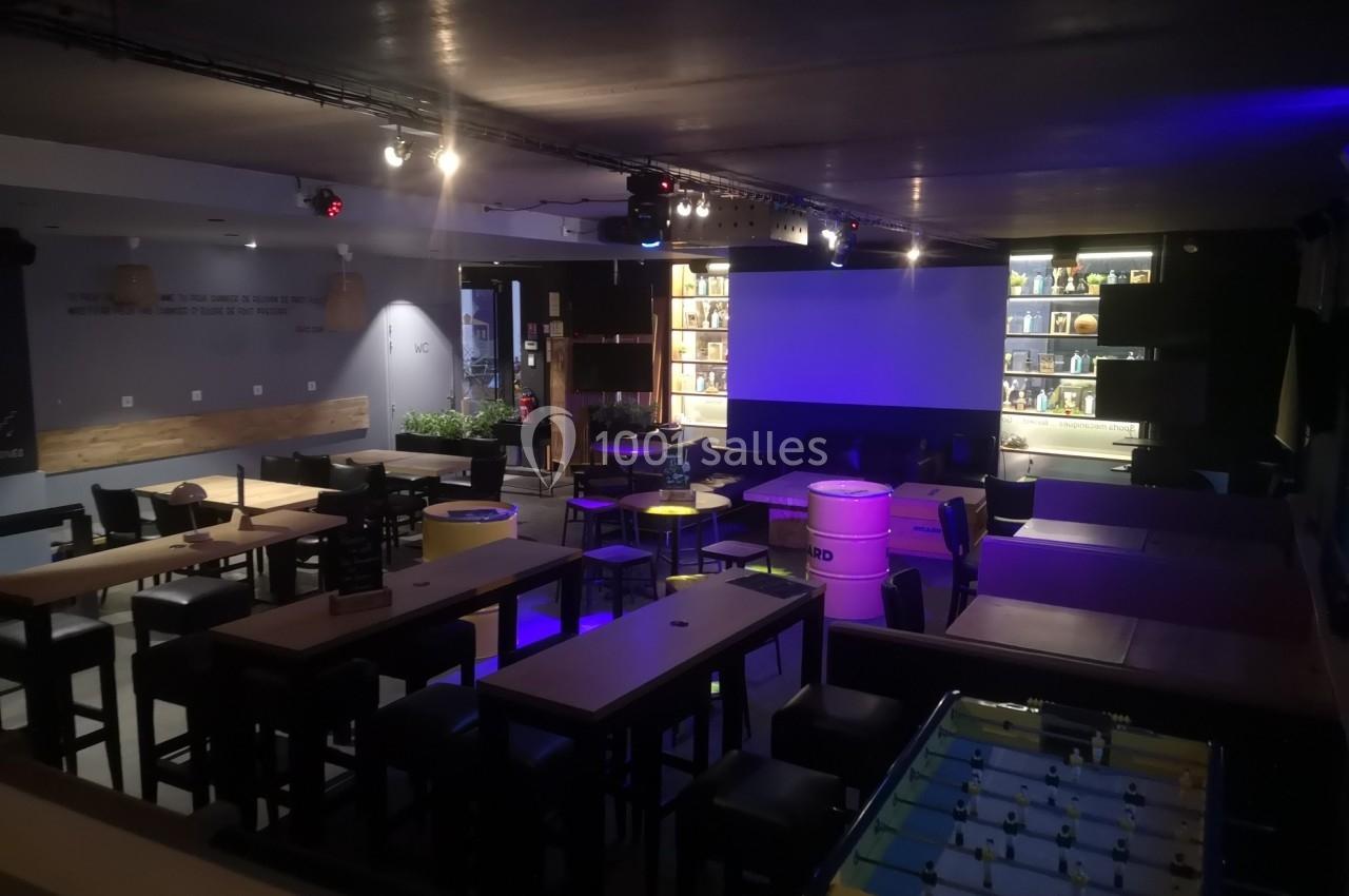 Salle sombre avec tables hautes, chaises, baby-foot et écran de projection, éclairée par des lumières tamisées.