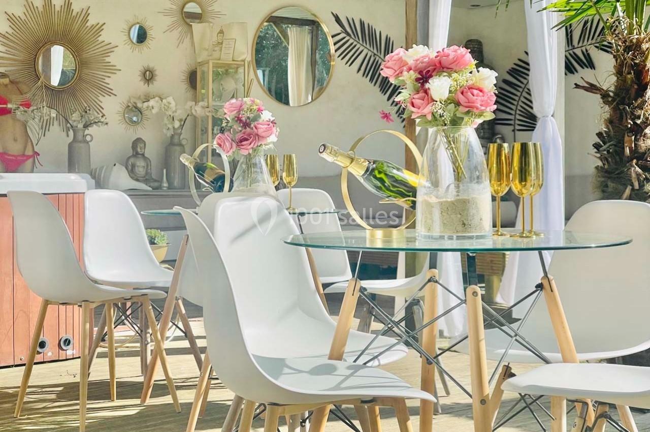 Table en verre entourée de chaises blanches, décorée de fleurs, verres dorés et bouteille de champagne, en extérieur.