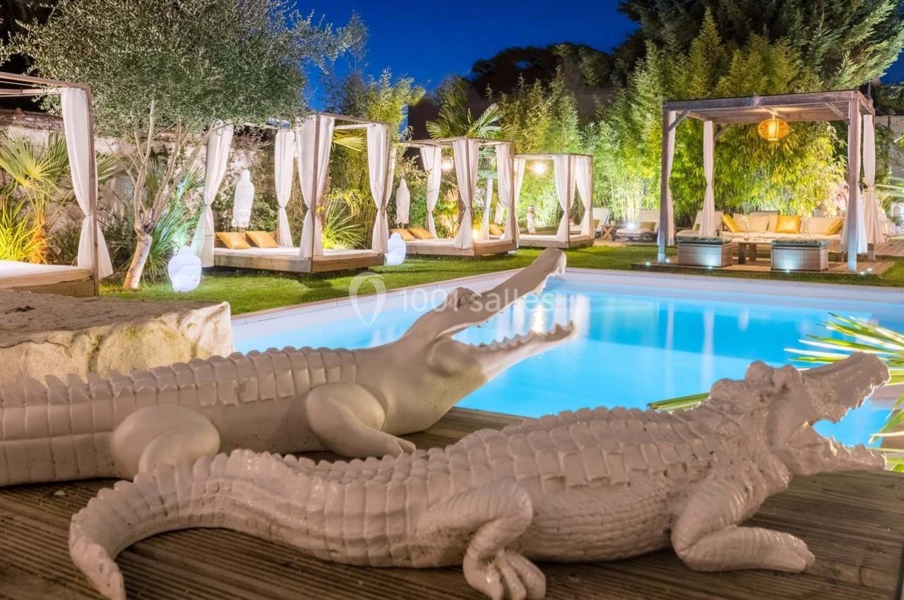 Statues de crocodiles blancs en céramique près d'une piscine éclairée, entourée de lits baldaquins et de végétation.