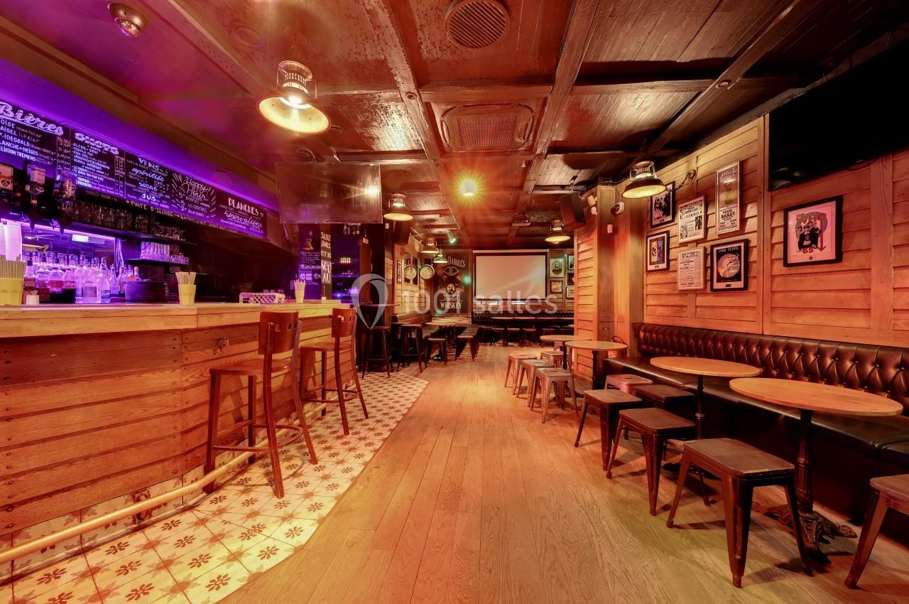 Intérieur d'un bar avec comptoir en bois, tabourets, tables basses, banquette et éclairage tamisé.