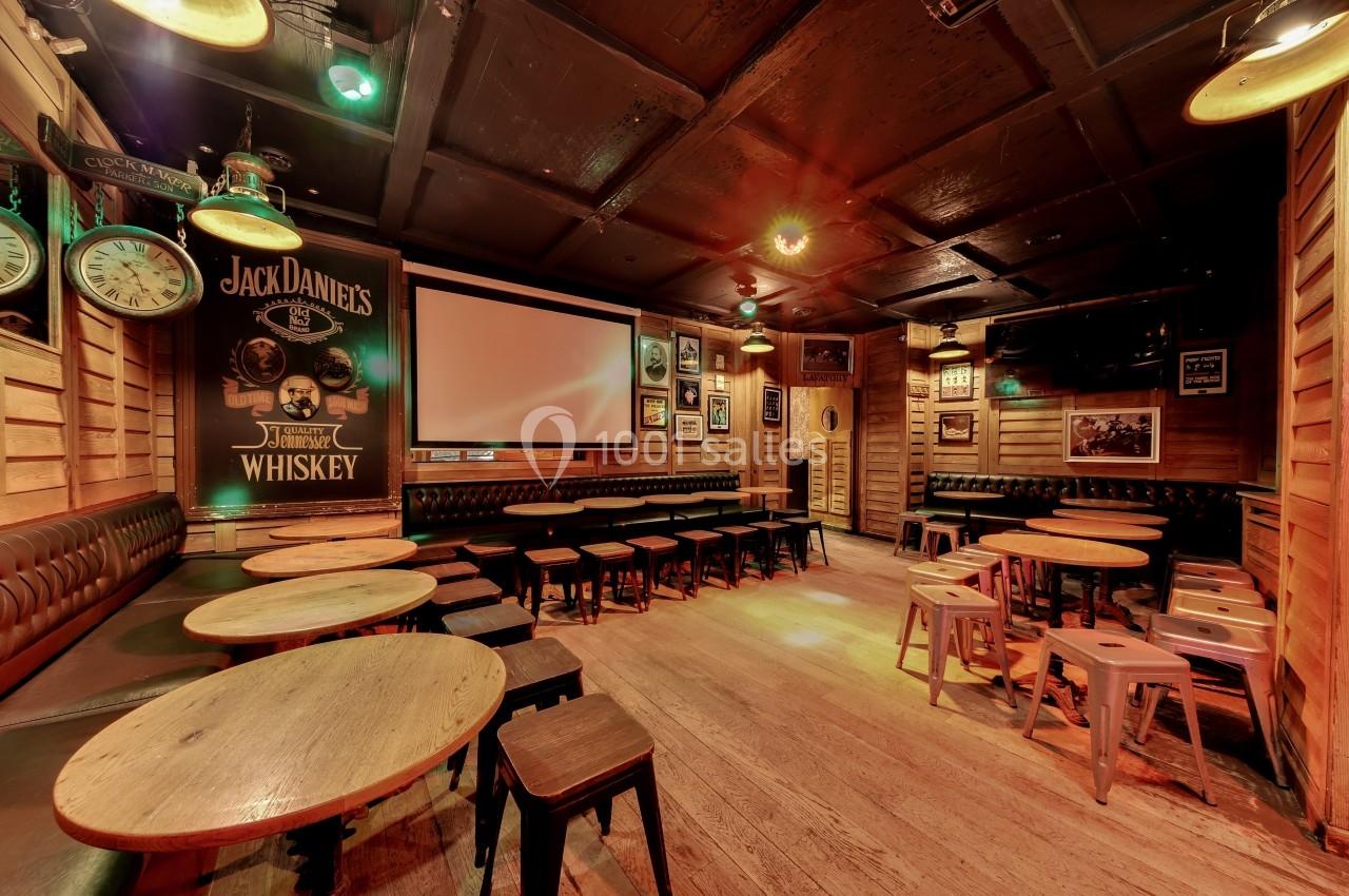 Salle de bar chaleureuse avec tables en bois, tabourets, banquettes et décorations murales rétro sous un éclairage tamisé.