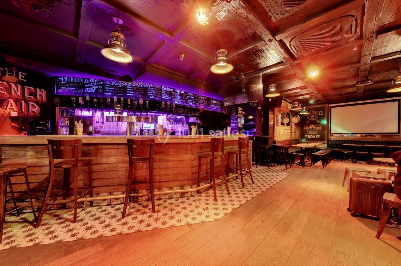 Intérieur d'un bar avec un long comptoir en bois, des tabourets alignés et un éclairage tamisé.