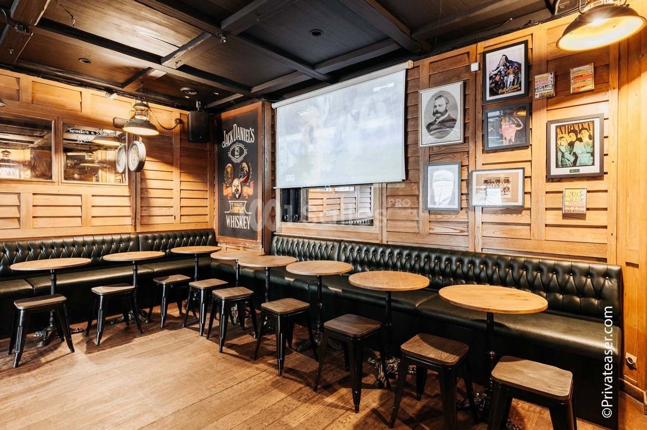 Intérieur d'un bar avec des tables rondes, des tabourets en bois et des murs décorés de cadres et affiches.