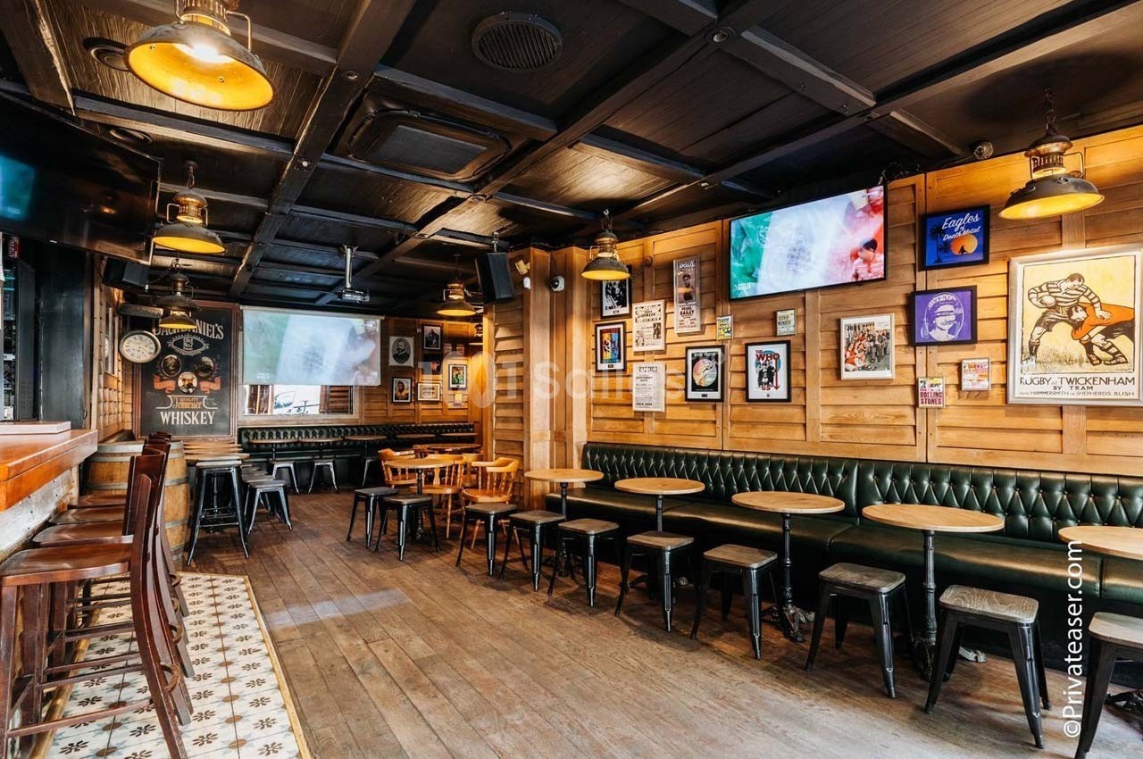 Salle de bar avec tables en bois, banquettes vertes, murs décorés d'affiches et écrans suspendus.