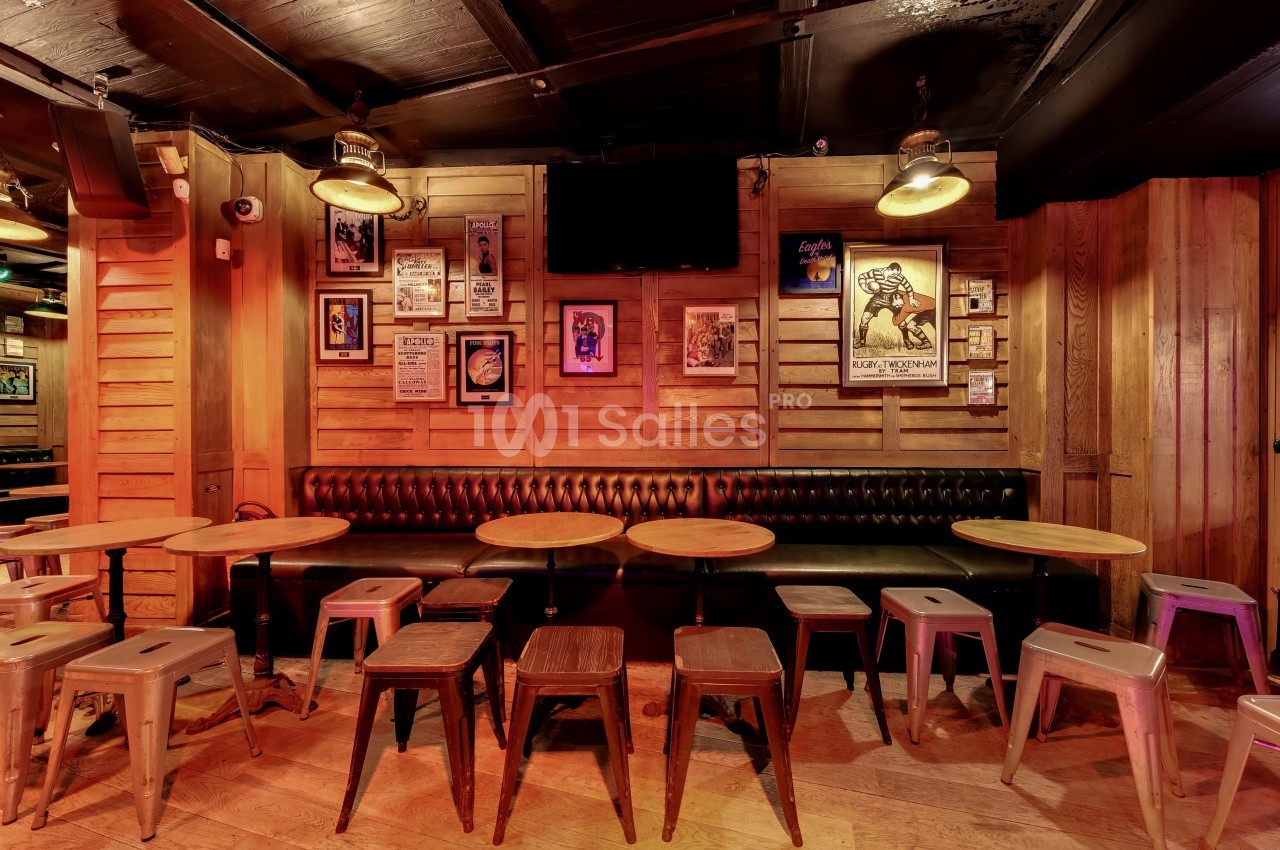 Salle de bar avec tables rondes, tabourets en métal et banquette en cuir, décorée de cadres sur des murs en bois.