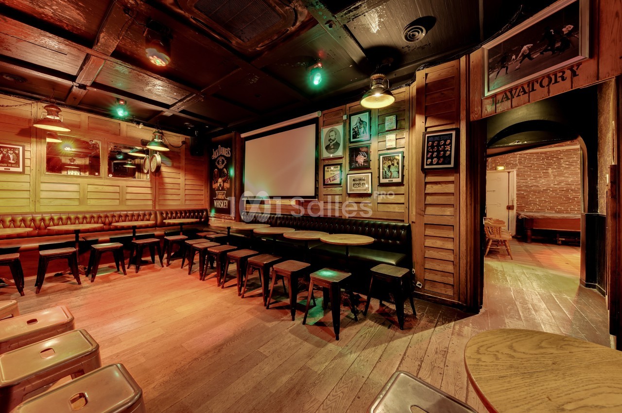 Salle de bar en bois avec tables, tabourets, écran mural et décorations vintage sur les murs.
