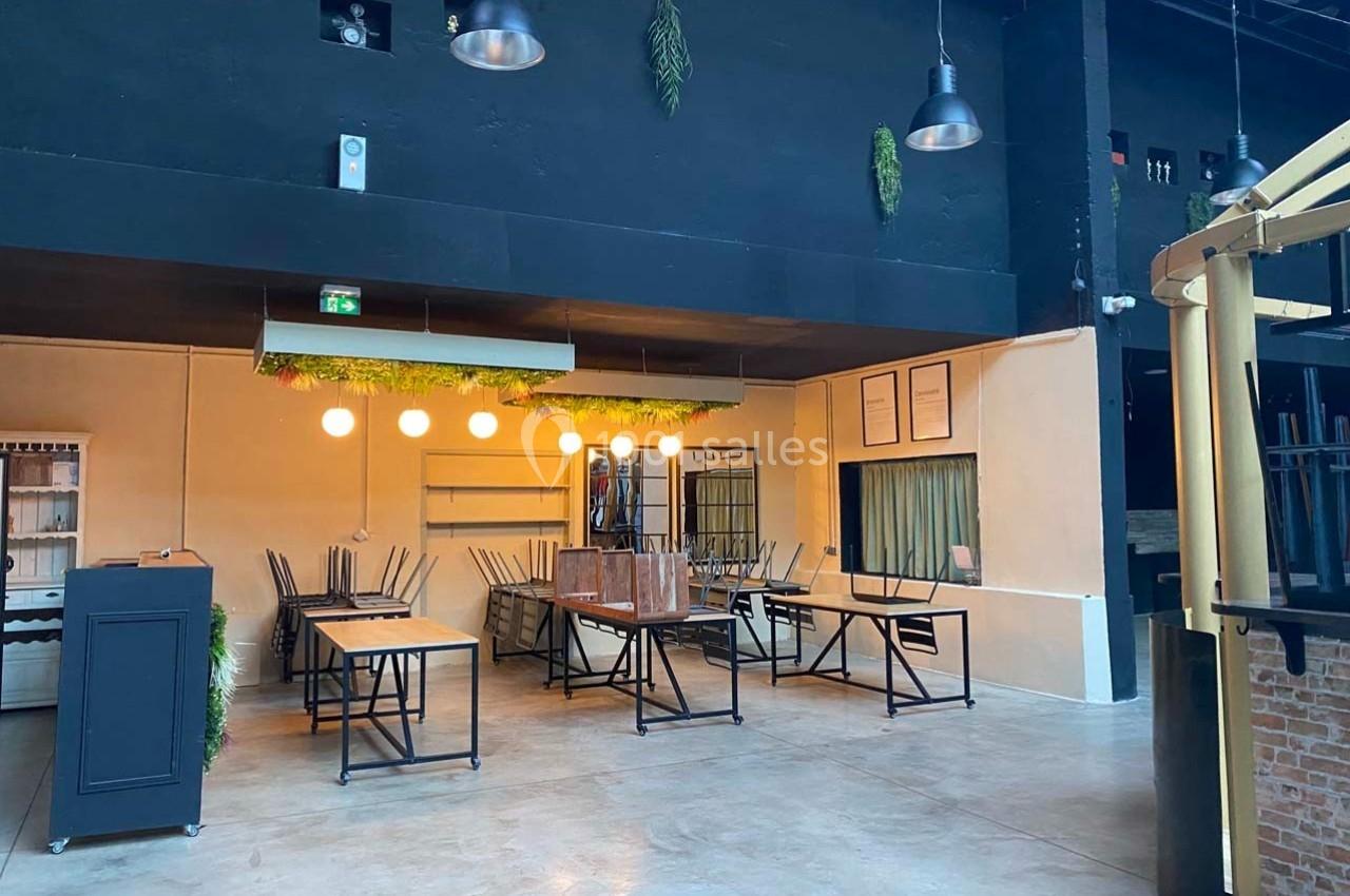 Salle vide avec tables et chaises pliées, éclairée par des suspensions lumineuses et décorée de touches végétales.