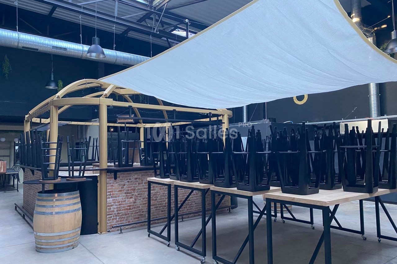 Chaises empilées sur des tables dans un espace intérieur avec un bar en briques et une grande toile tendue au plafond.