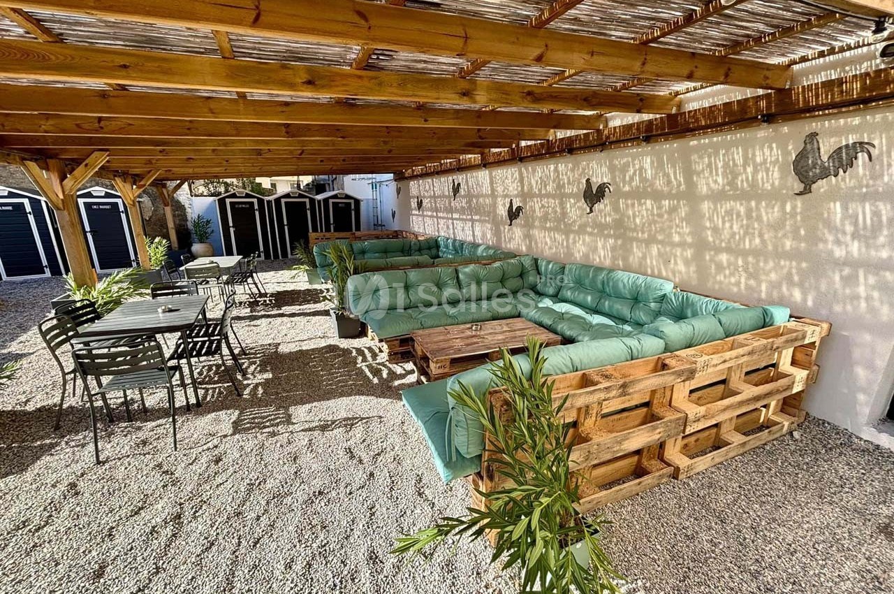 Terrasse ombragée avec salon en palettes et coussins verts, tables en métal noir, sol en gravier et décor mural.