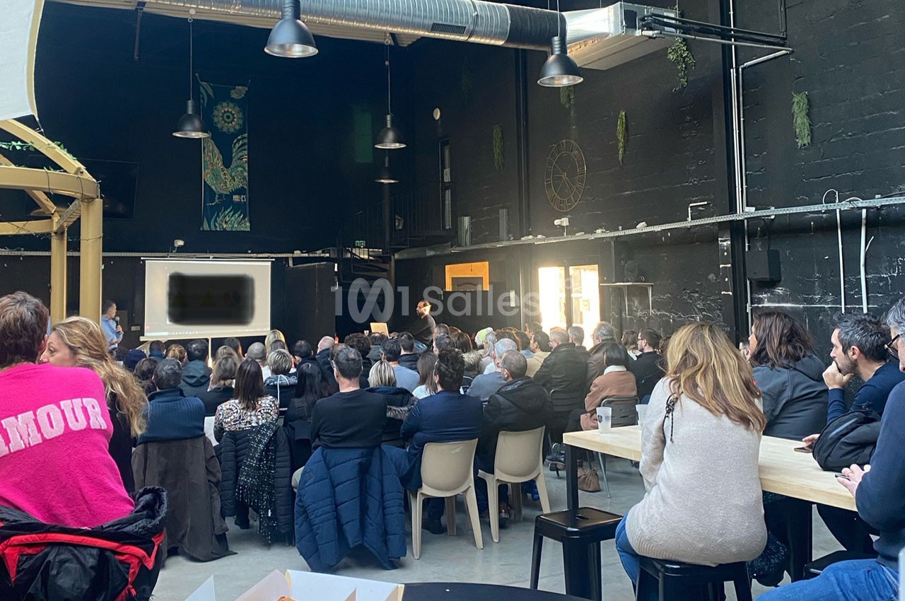 Un groupe de personnes assiste à une présentation dans une grande salle sombre avec des tables et des chaises.