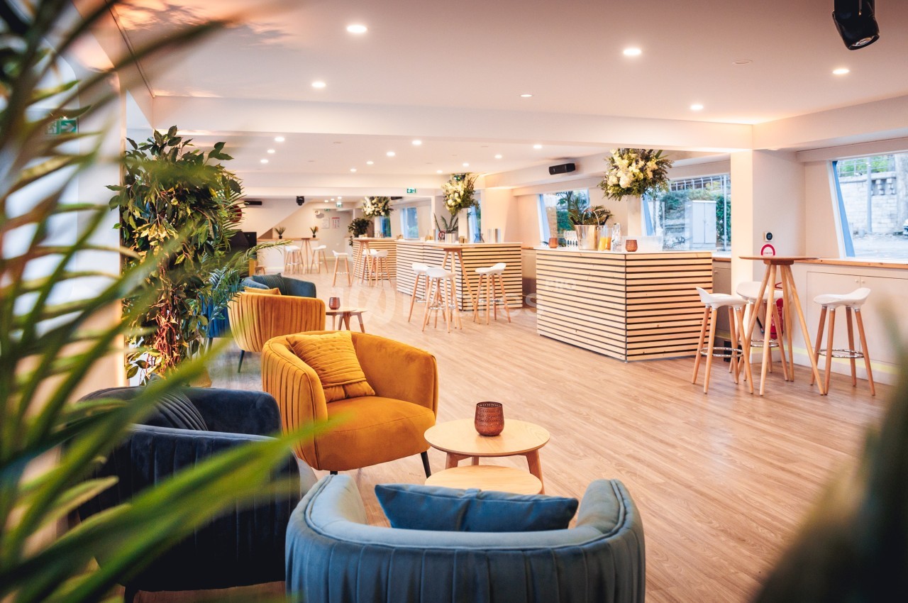 Salle lumineuse avec fauteuils colorés, tables basses, bar en bois clair et plantes décoratives.