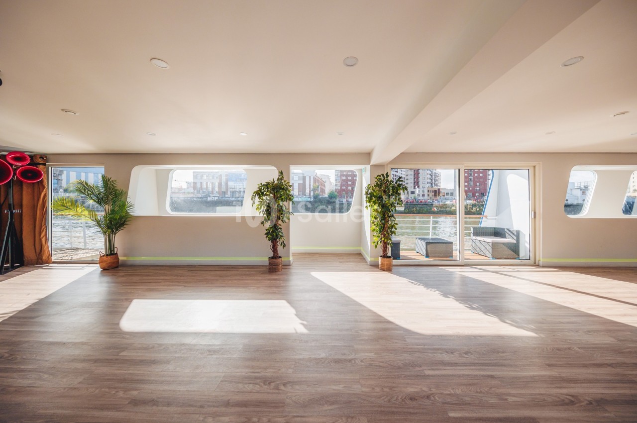 Salle lumineuse avec parquet, grandes fenêtres donnant sur l'extérieur, plantes en pot et vue sur des bâtiments.