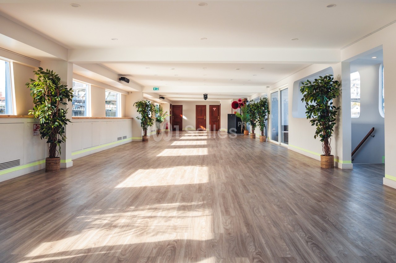 Salle lumineuse avec parquet en bois, grandes fenêtres laissant entrer la lumière et plantes disposées le long des murs.