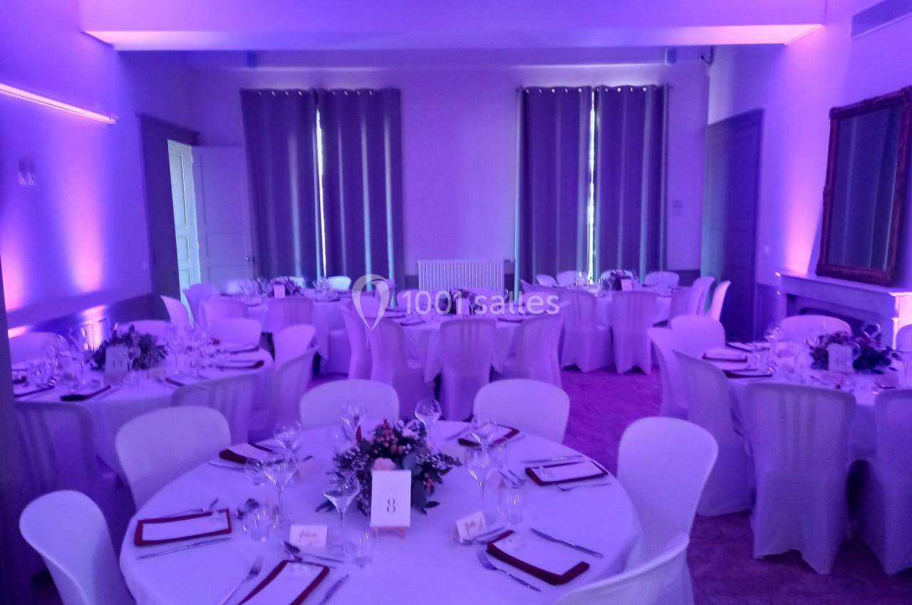 Salle de réception éclairée en violet, avec des tables rondes dressées, nappes blanches et décorations florales.