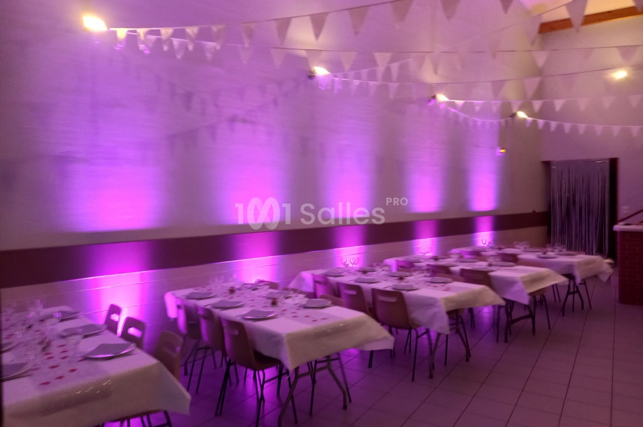 Salle décorée pour un événement, avec tables dressées, nappes blanches et éclairage d'ambiance rose.