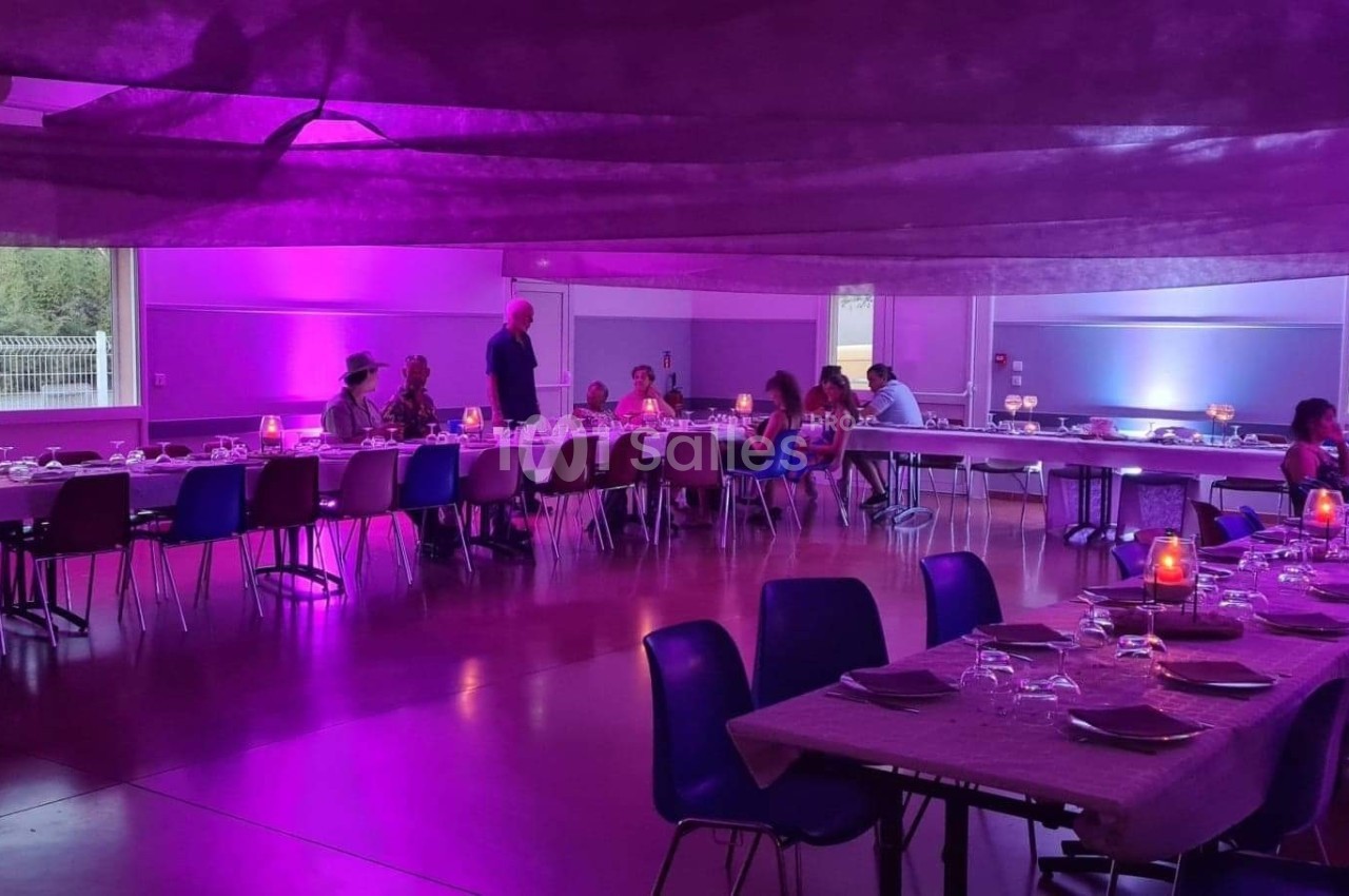 Salle décorée avec des tables dressées et éclairage violet, des convives assis et debout discutant.
