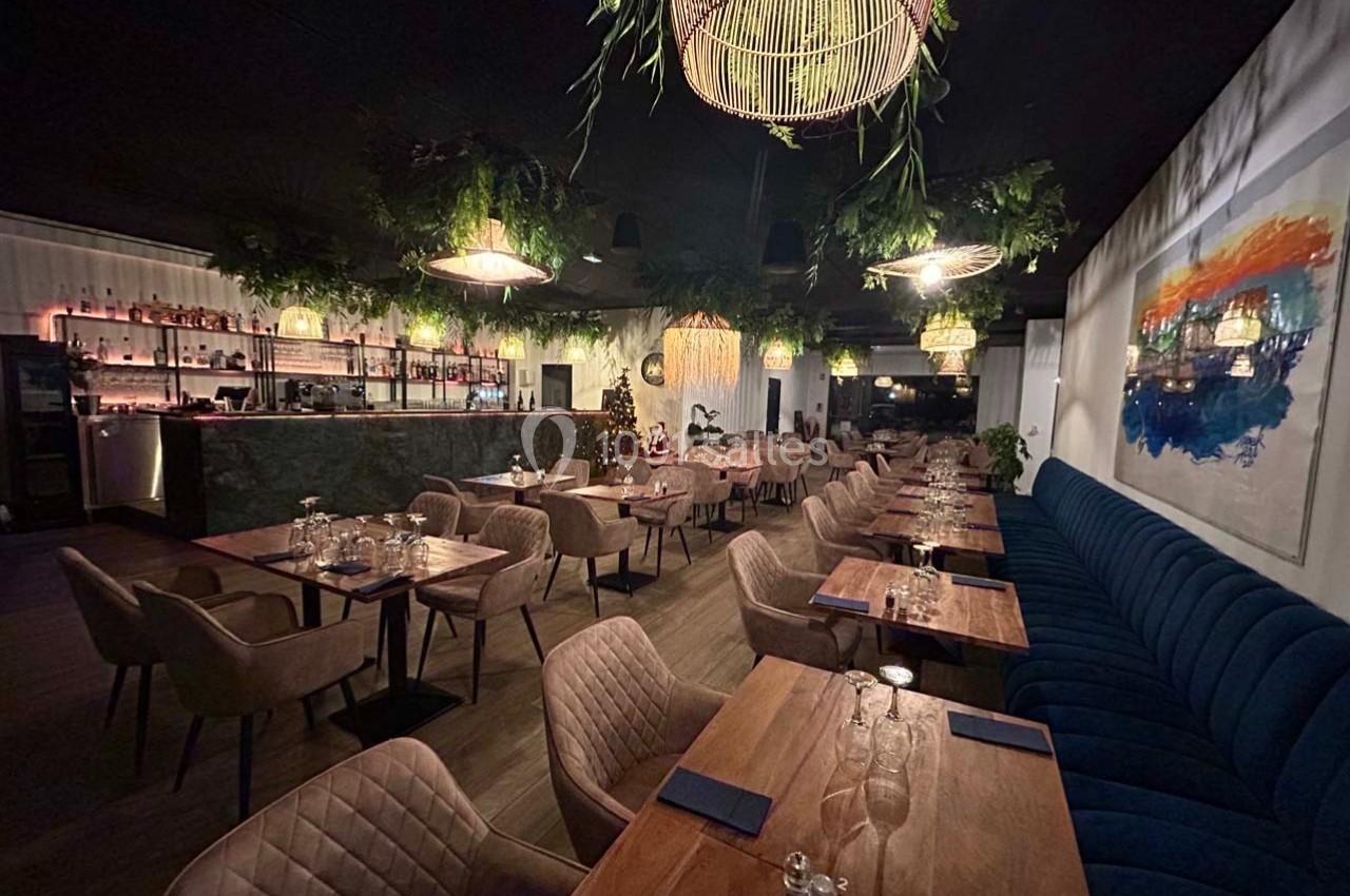 Salle de restaurant élégante avec tables en bois, chaises rembourrées, bar éclairé et décoration végétale suspendue.