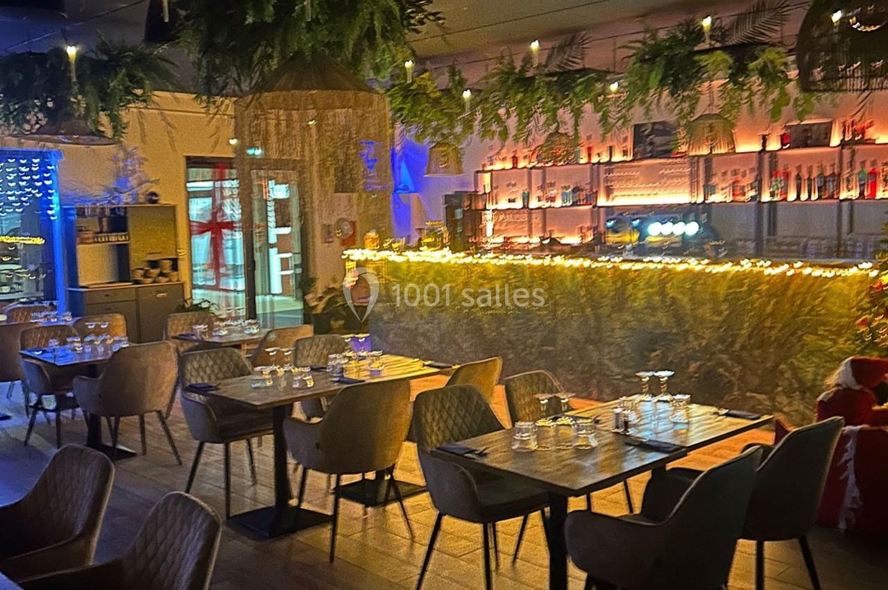 Salle de restaurant décorée avec des guirlandes lumineuses, des plantes suspendues et un bar éclairé en arrière-plan.