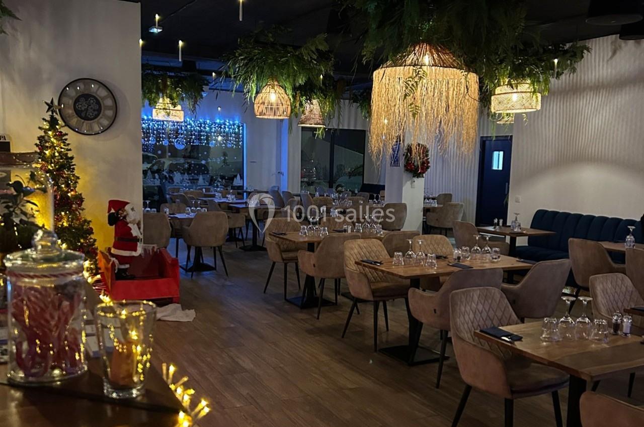 Salle de restaurant décorée pour Noël avec des tables dressées, des guirlandes lumineuses et un sapin en arrière-plan.