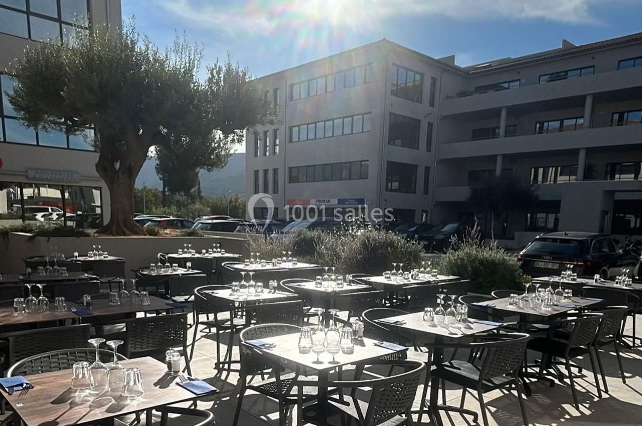 Terrasse d'un restaurant avec des tables dressées, entourée de bâtiments modernes et éclairée par le soleil.