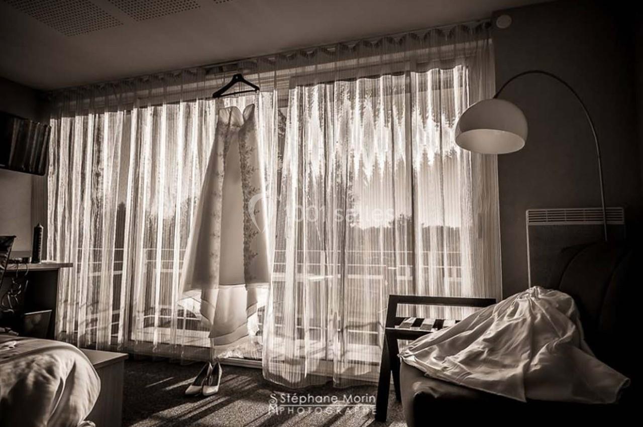 Robe de mariée suspendue devant une baie vitrée laissant passer la lumière dans une chambre.