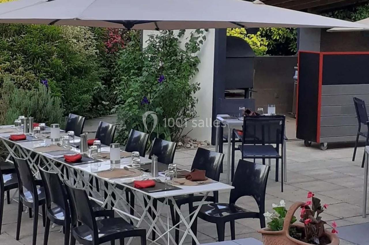 Terrasse aménagée avec tables dressées, chaises noires, parasol et barbecue en arrière-plan, entourée de verdure.