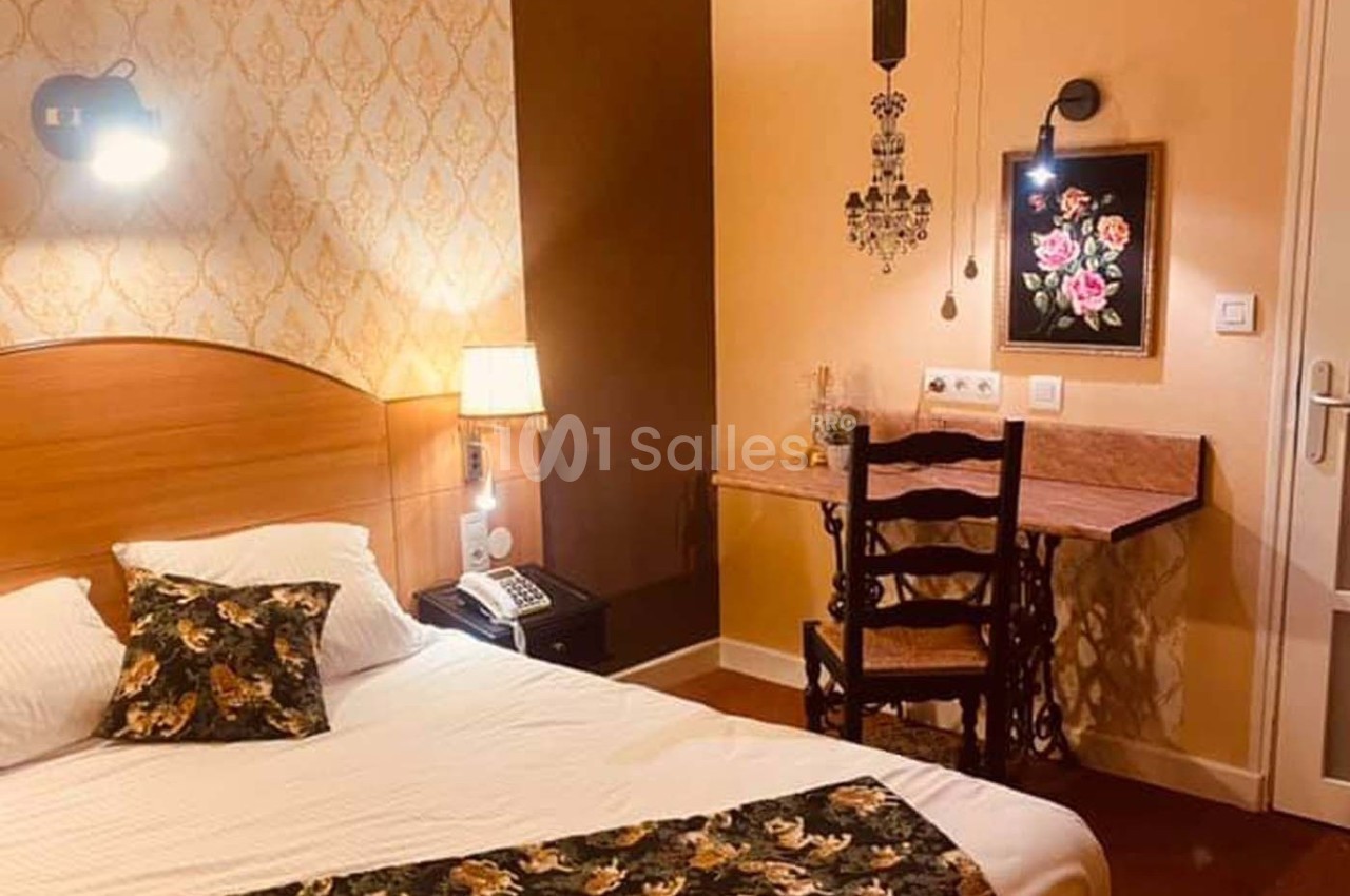 Chambre avec lit double, bureau en bois, chaise ancienne, éclairage mural et décoration florale sur un mur beige.