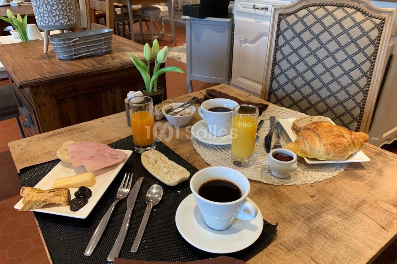 Petit-déjeuner servi sur une table avec café, jus d'orange, croissant, pain, confiture, jambon et décor rustique.
