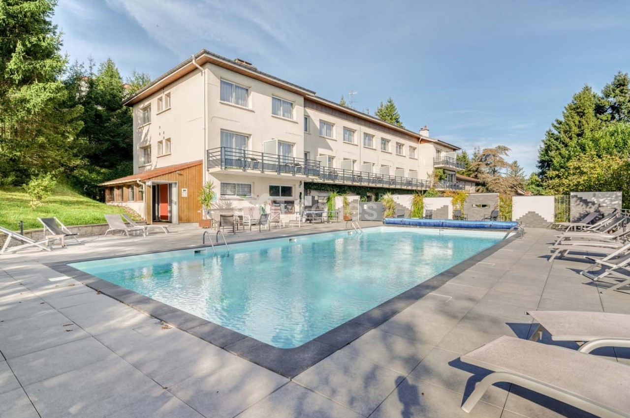 Piscine extérieure entourée de chaises longues, située devant un bâtiment résidentiel avec un jardin arboré.