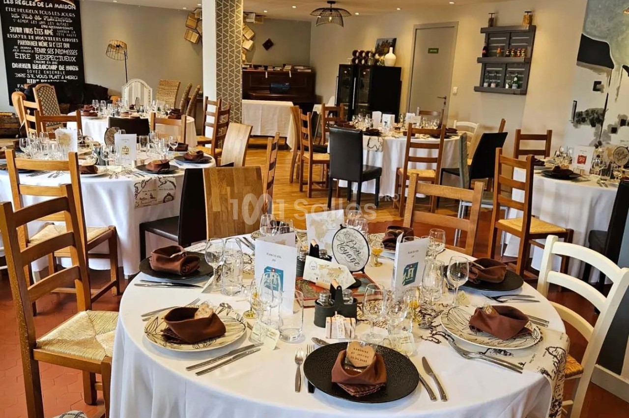 Salle de restaurant décorée avec des tables rondes dressées, nappes blanches, serviettes marron et chaises en bois.
