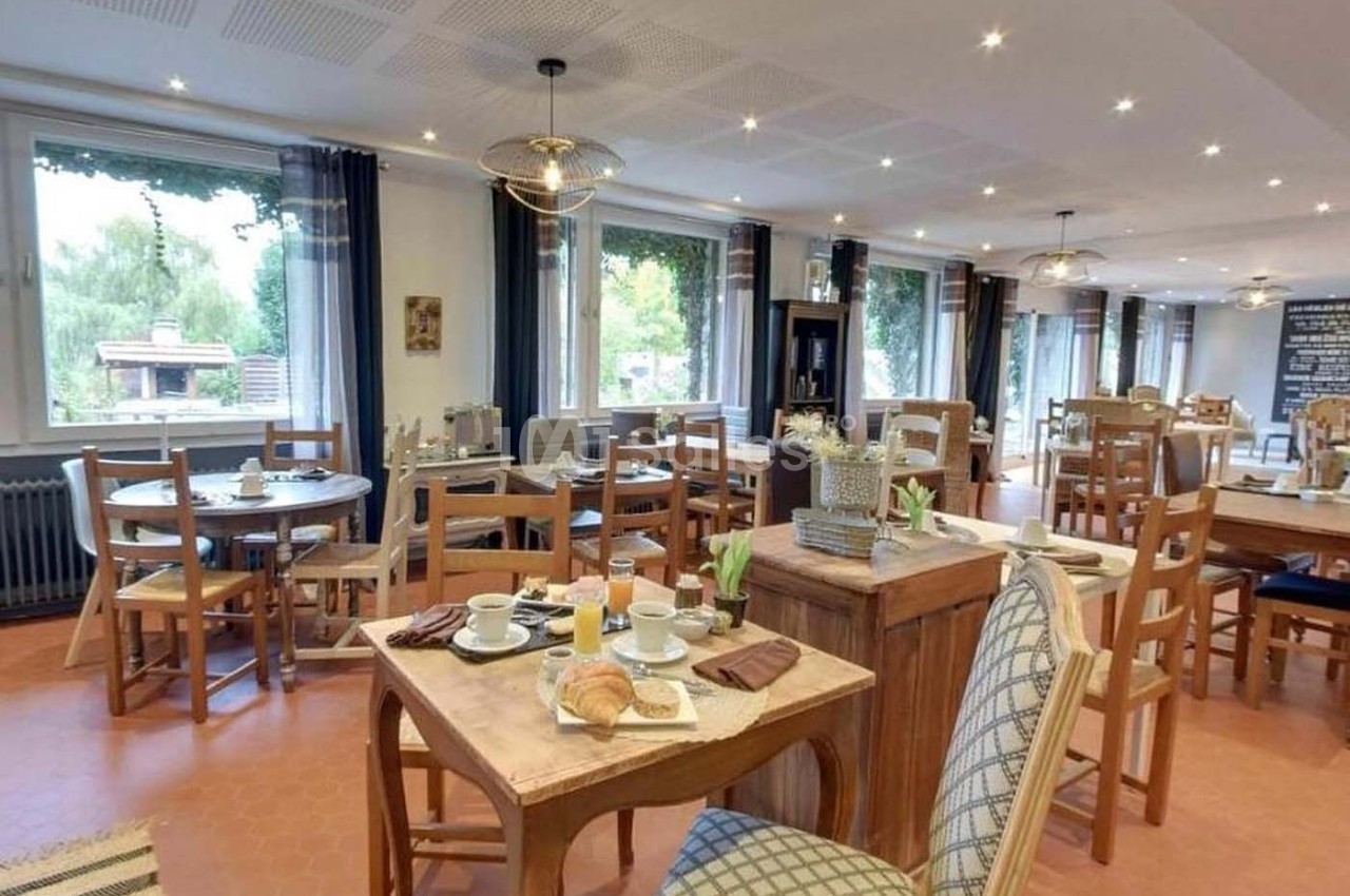 Salle lumineuse avec tables en bois dressées pour le petit-déjeuner, grandes fenêtres et décoration chaleureuse.