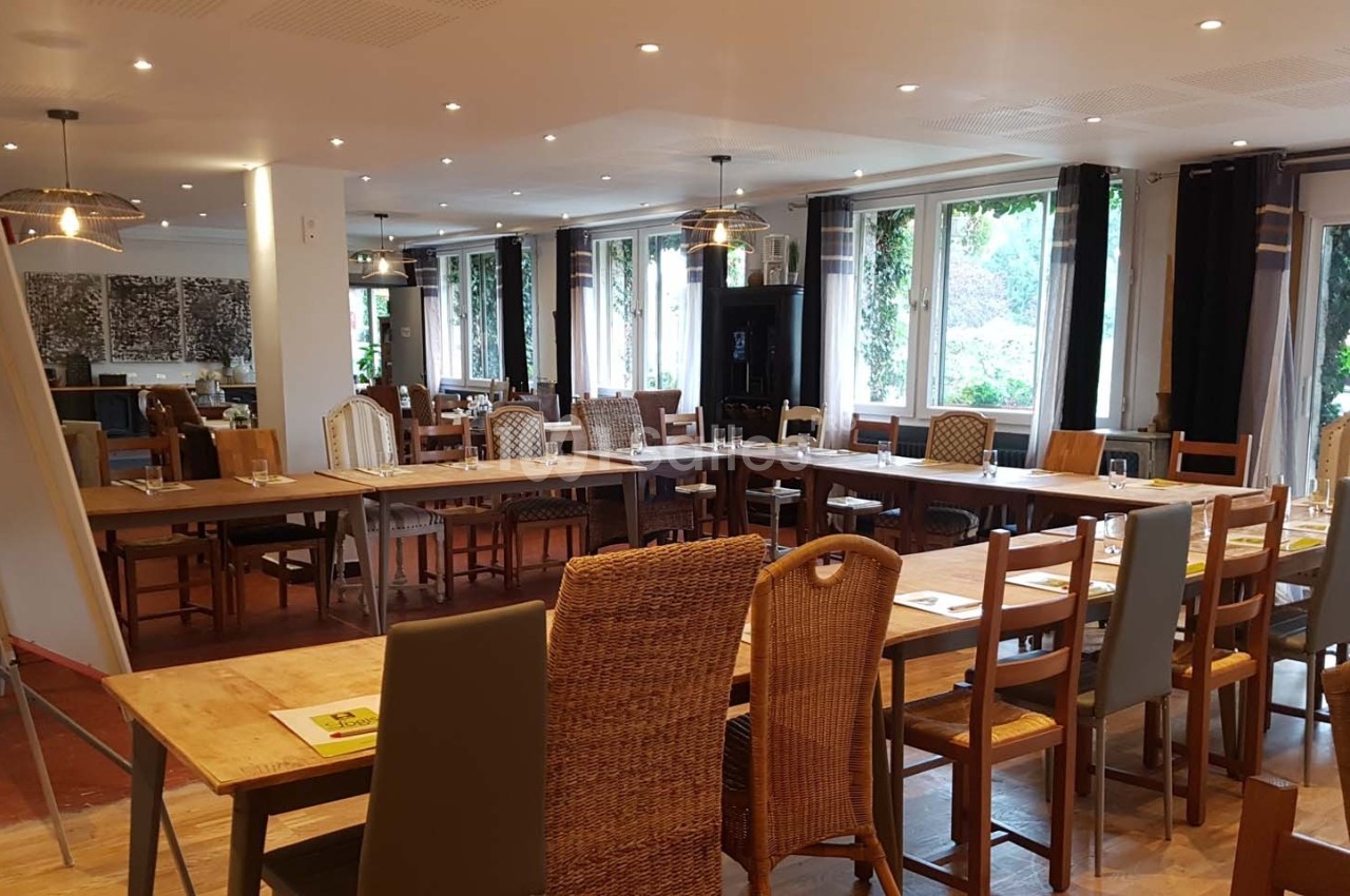 Salle lumineuse aménagée avec de longues tables en bois et des chaises variées, entourée de grandes fenêtres.
