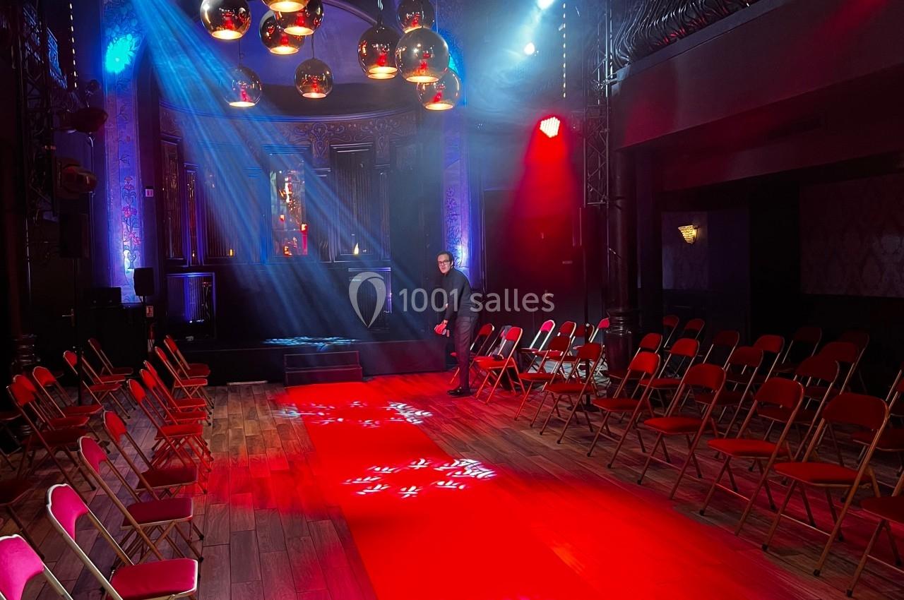 Salle de spectacle avec tapis rouge central, chaises alignées, éclairages colorés et décorations suspendues.