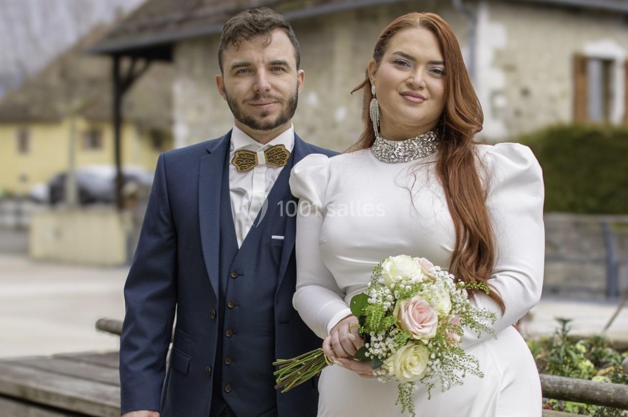 Un couple en tenue de mariage pose devant un bâtiment en pierre avec un bouquet de fleurs.
