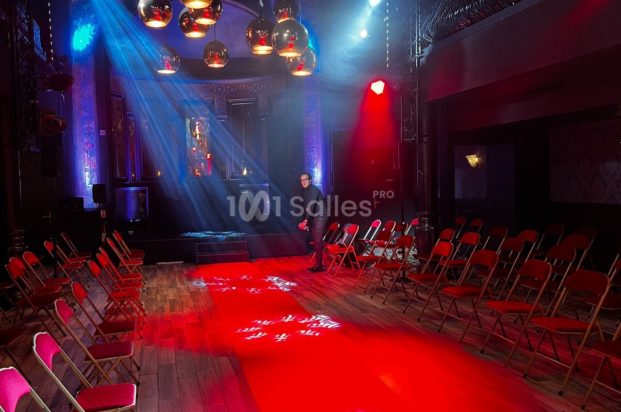 Salle sombre avec un tapis rouge central, rangées de chaises pliantes rouges et éclairage de scène coloré.