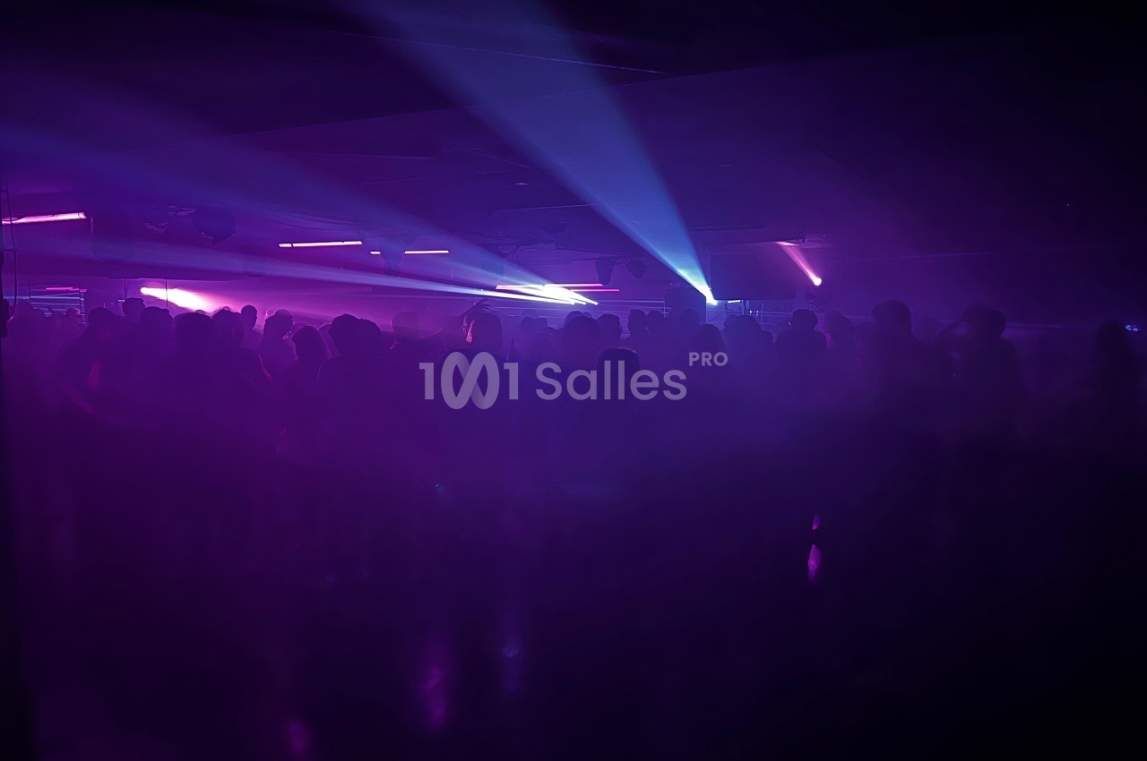 Foule dans une salle sombre éclairée par des lumières violettes et bleues, créant une ambiance festive.