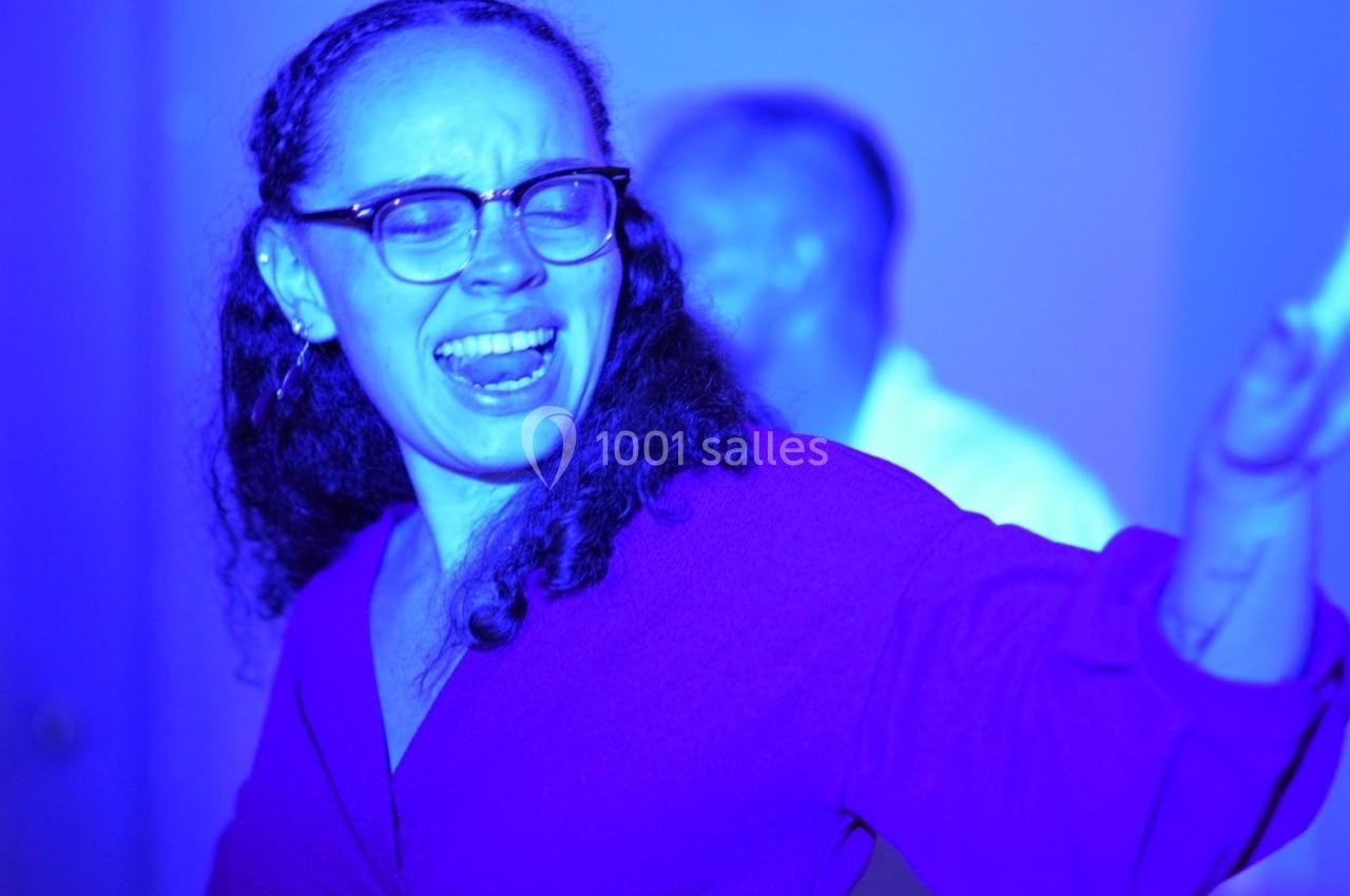 Femme portant des lunettes, chantant ou s'exprimant avec énergie dans une lumière bleue vive.