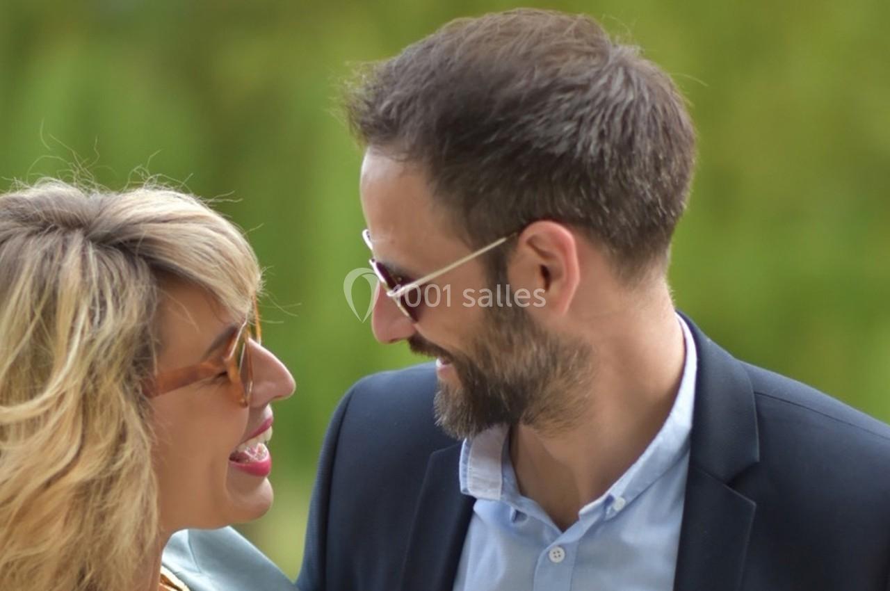 Un homme et une femme sourient en se regardant, portant des lunettes de soleil, en extérieur avec un fond verdoyant.