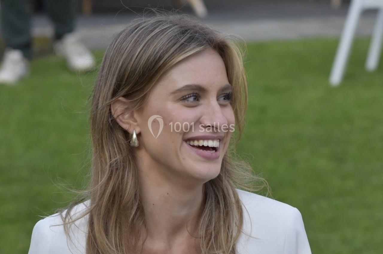 Femme souriante en extérieur, portant une veste blanche, avec une pelouse verte en arrière-plan.